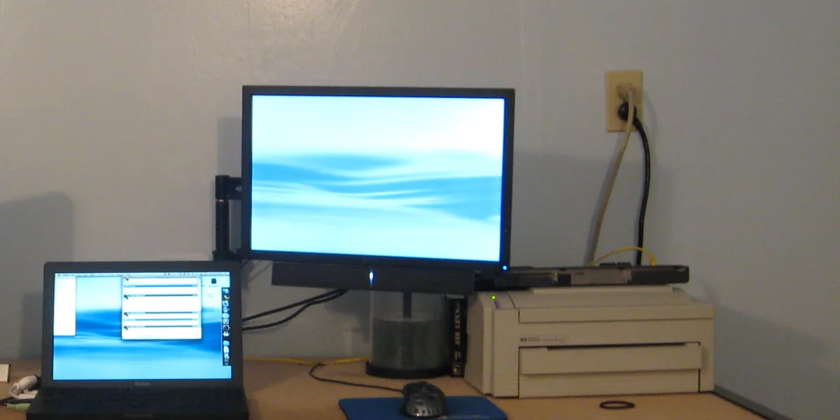 Monitor an einer Halterung mit Säule über einer einfachen Tischplatte, links davor ein schwarzer Laptop, rechts ein heller Drucker, an der Wand eine Steckdose