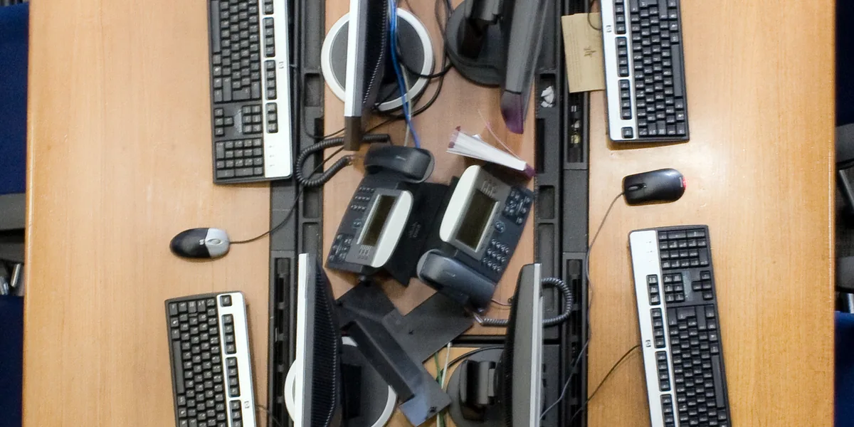 Holztisch von oben mit vier Tastaturen, zwei Mäusen, Telefonen und den Oberkanten mehrerer Monitore, in der Mitte zwei Kabelkanäle
