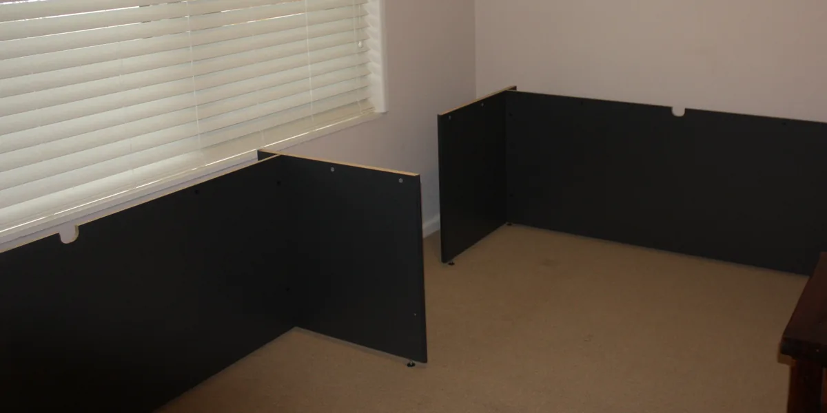 Schwarze Seitenteile und Rückwände zweier Schreibtische ohne Platten in einem Zimmer mit Teppichboden, links ein Fenster mit heller Jalousie